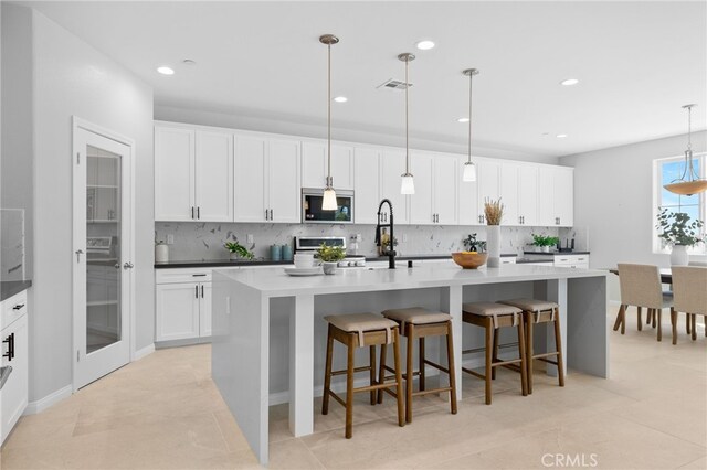 kitchen with an island with sink, backsplash, stainless steel microwave, and white cabinetry