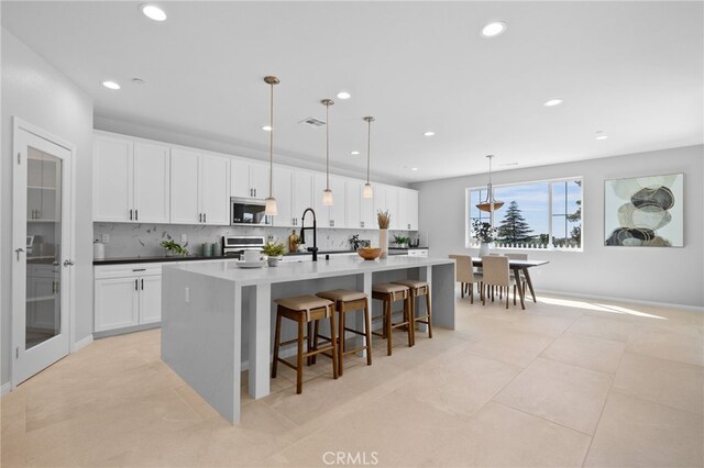 kitchen with recessed lighting, an island with sink, backsplash, stainless steel microwave, and white cabinetry