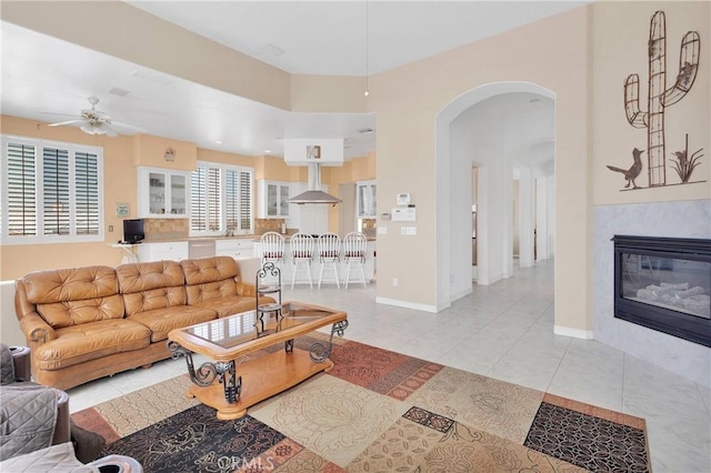 living room featuring a ceiling fan, arched walkways, light tile patterned flooring, baseboards, and a glass covered fireplace