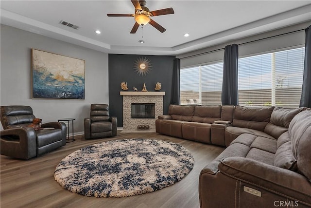 living area featuring wood finished floors, a glass covered fireplace, ceiling fan, a tray ceiling, and visible vents