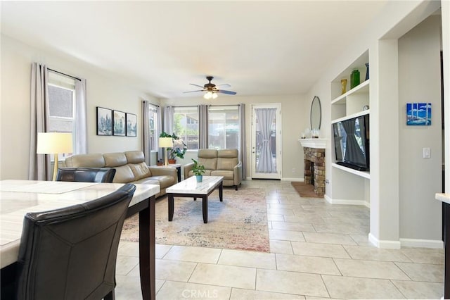 living area with light tile patterned flooring, a stone fireplace, built in shelves, a ceiling fan, and baseboards