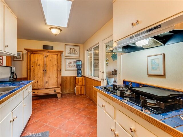 kitchen with a skylight, tile countertops, a wainscoted wall, under cabinet range hood, and black gas cooktop