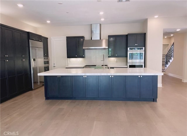 kitchen featuring wall chimney range hood, stainless steel appliances, light wood-type flooring, and light countertops