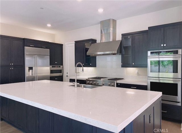 kitchen featuring glass insert cabinets, wall chimney range hood, a large island with sink, a sink, and appliances with stainless steel finishes