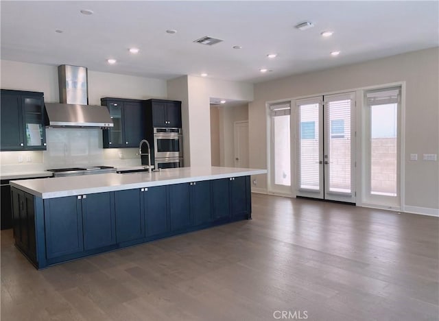 kitchen featuring wall chimney range hood, a spacious island, wood finished floors, light countertops, and double oven