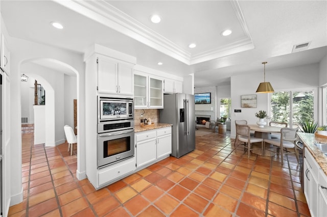 kitchen featuring stainless steel appliances, a raised ceiling, decorative backsplash, crown molding, and a large fireplace