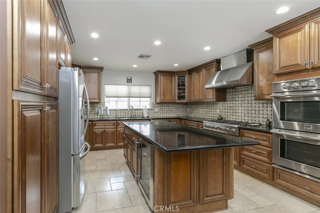 kitchen with wall chimney range hood, brown cabinets, appliances with stainless steel finishes, beverage cooler, and backsplash