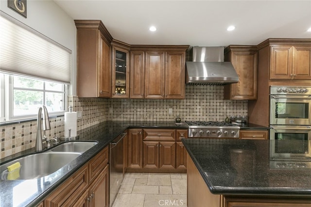 kitchen featuring brown cabinetry, stone tile floors, wall chimney range hood, a sink, and stainless steel appliances