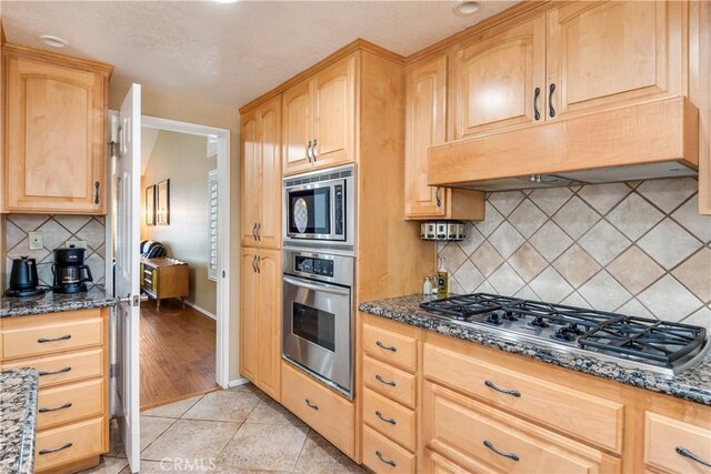 kitchen featuring light brown cabinetry, dark stone counters, stainless steel appliances, and under cabinet range hood
