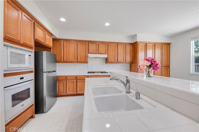 kitchen featuring a sink, under cabinet range hood, brown cabinetry, tile countertops, and white appliances