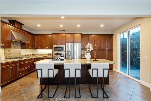 kitchen with range hood, stainless steel appliances, a center island with sink, a breakfast bar, and backsplash
