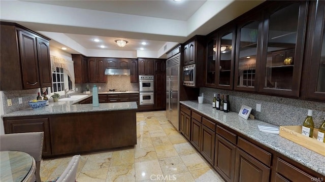 kitchen featuring tasteful backsplash, dark brown cabinetry, under cabinet range hood, built in appliances, and a sink