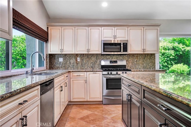 kitchen with a sink, backsplash, stone countertops, cream cabinetry, and appliances with stainless steel finishes