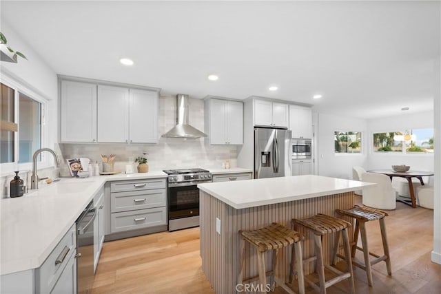 kitchen featuring gray cabinets, a sink, wall chimney exhaust hood, a kitchen bar, and appliances with stainless steel finishes