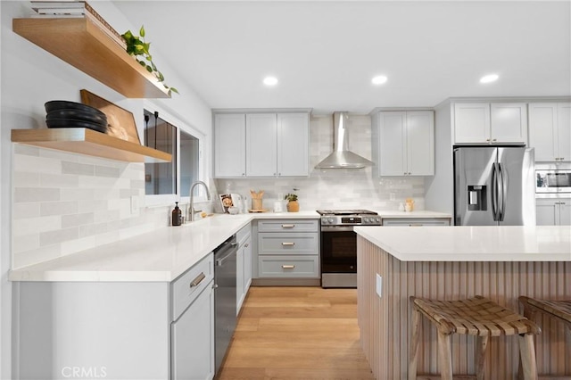 kitchen featuring a sink, appliances with stainless steel finishes, wall chimney range hood, open shelves, and a breakfast bar area