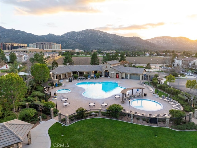 pool at dusk featuring a yard, a mountain view, a community pool, and fence