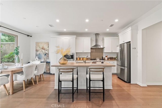 kitchen with wall chimney range hood, white cabinets, stainless steel appliances, light countertops, and a center island with sink