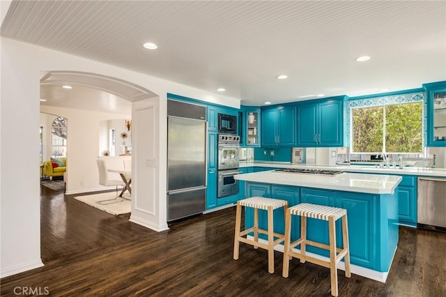 kitchen featuring blue cabinets, arched walkways, light countertops, built in appliances, and dark wood-style floors