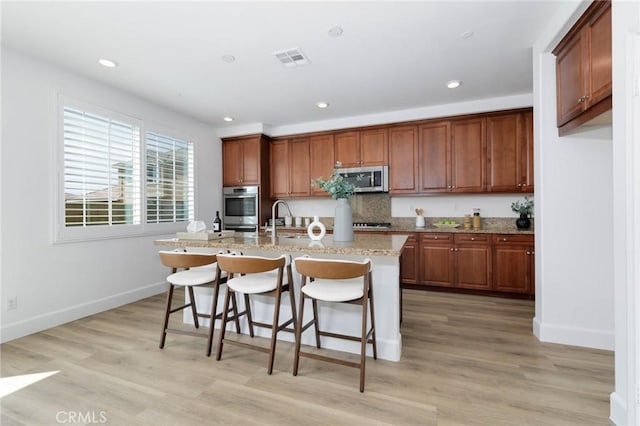 kitchen with a kitchen breakfast bar, brown cabinetry, visible vents, stainless steel microwave, and an island with sink