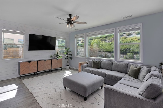 living room with wood walls, light wood finished floors, baseboards, visible vents, and a ceiling fan