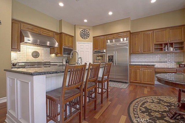 kitchen featuring stainless steel appliances, under cabinet range hood, dark wood-type flooring, and decorative backsplash