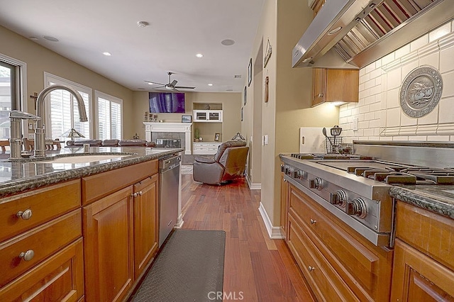 kitchen with exhaust hood, brown cabinets, stainless steel appliances, a fireplace, and ceiling fan