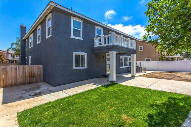 rear view of property with fence, a balcony, a patio, stucco siding, and a lawn