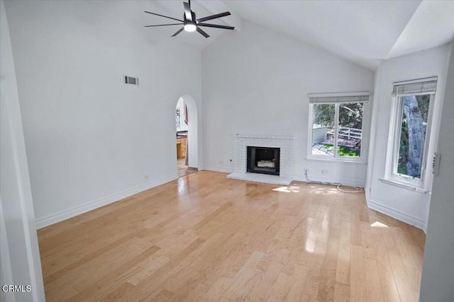 unfurnished living room featuring a brick fireplace, ceiling fan, high vaulted ceiling, light wood-style flooring, and arched walkways