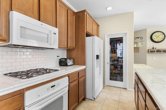 kitchen with light tile patterned floors, brown cabinets, decorative backsplash, tile counters, and white appliances