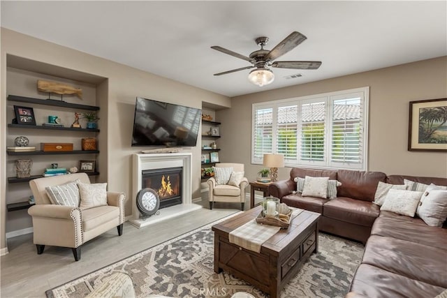 living area with visible vents, baseboards, a glass covered fireplace, light wood finished floors, and a ceiling fan