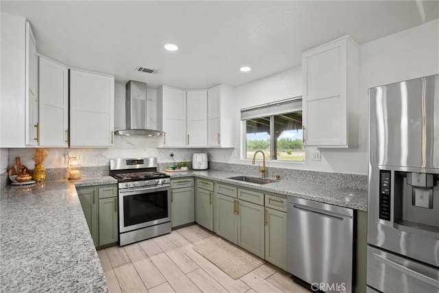 kitchen with green cabinetry, white cabinets, a sink, appliances with stainless steel finishes, and wall chimney exhaust hood