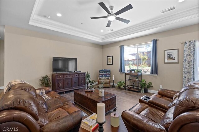 living room with a raised ceiling, visible vents, ornamental molding, wood finished floors, and a ceiling fan