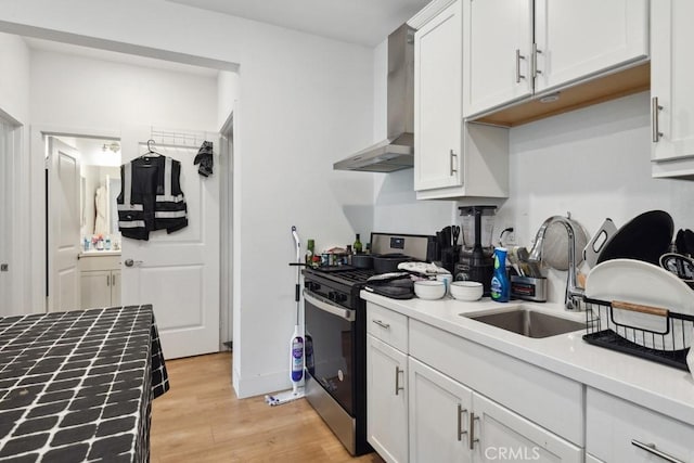 kitchen with wall chimney range hood, white cabinets, a sink, stainless steel gas stove, and light wood-style flooring