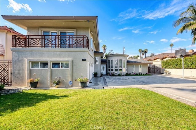 back of property featuring concrete driveway, a balcony, a gate, a lawn, and stucco siding