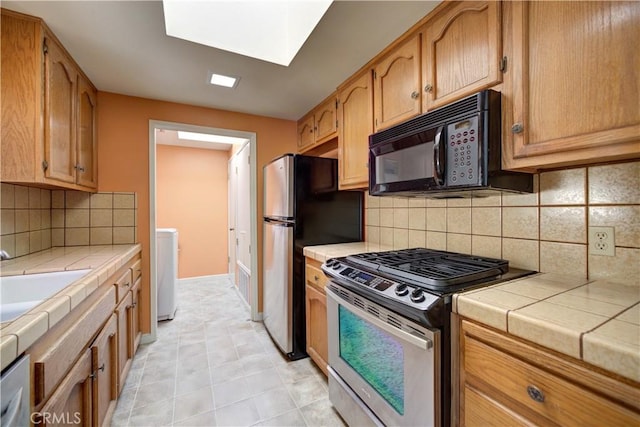 kitchen featuring appliances with stainless steel finishes, a sink, tile counters, baseboards, and a skylight
