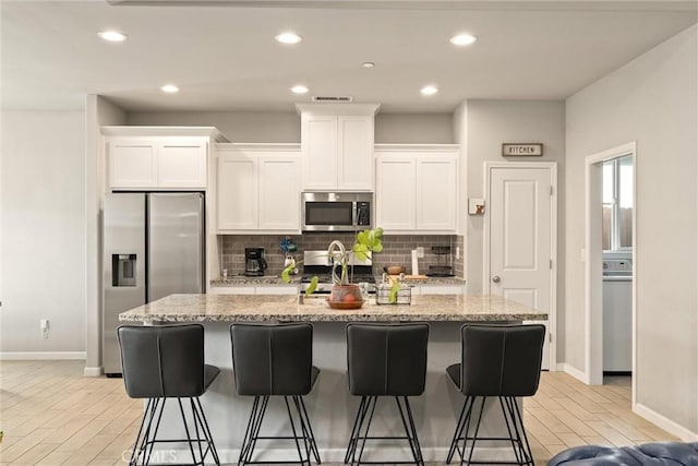 kitchen with a kitchen island with sink, white cabinetry, appliances with stainless steel finishes, backsplash, and baseboards