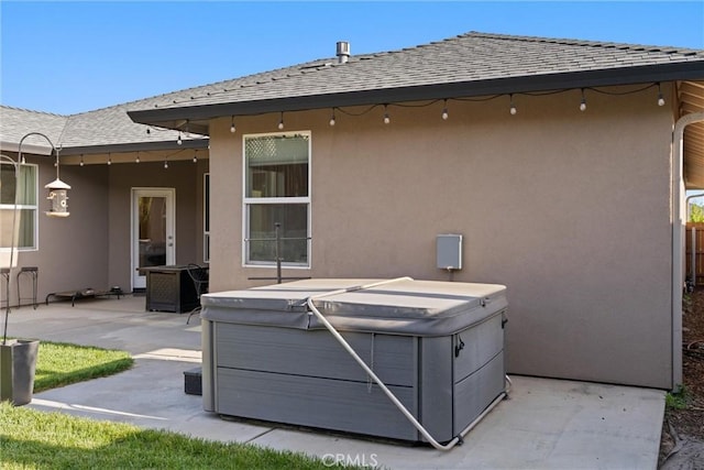rear view of house with a patio, stucco siding, a hot tub, and roof with shingles
