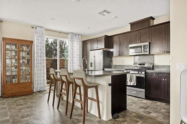 kitchen with dark brown cabinetry, an island with sink, stainless steel appliances, visible vents, and a kitchen breakfast bar