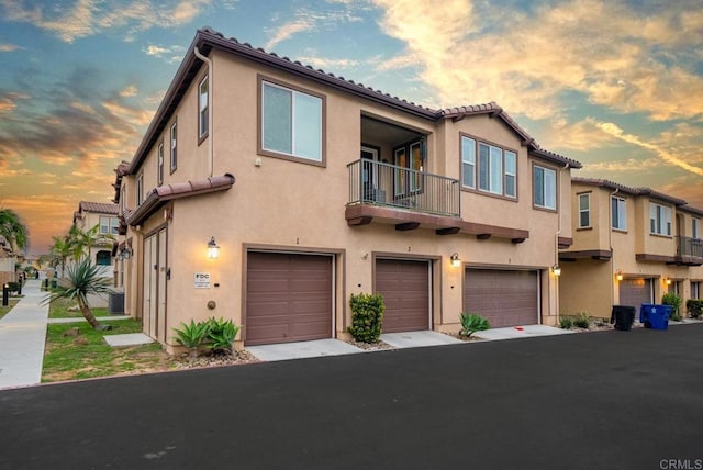 townhome / multi-family property featuring stucco siding, central AC unit, a tile roof, a balcony, and an attached garage