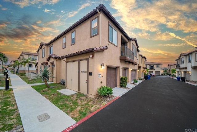 property exterior at dusk with a residential view, a garage, stucco siding, and a balcony