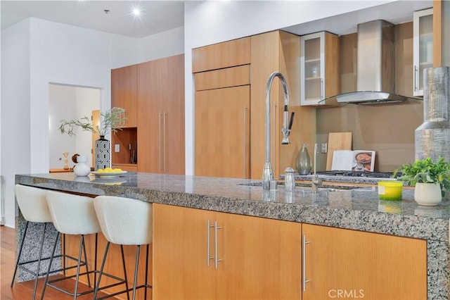 kitchen with glass insert cabinets, a sink, wall chimney exhaust hood, stone counters, and wood finished floors
