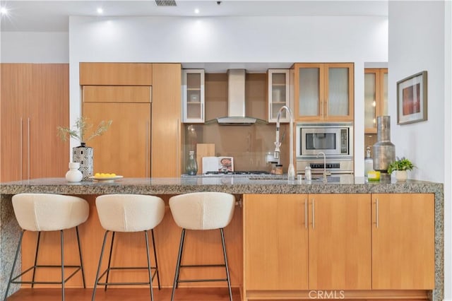 kitchen with stainless steel microwave, glass insert cabinets, wall chimney exhaust hood, a breakfast bar area, and stone countertops