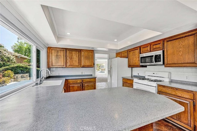 kitchen with a sink, a tray ceiling, a peninsula, backsplash, and white appliances