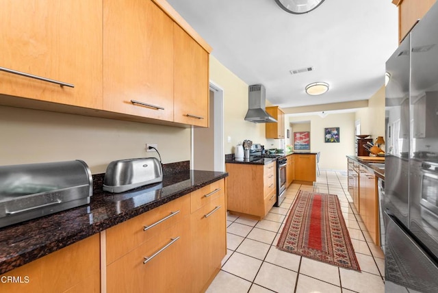 kitchen featuring wall chimney range hood, light tile patterned flooring, appliances with stainless steel finishes, dark stone counters, and visible vents