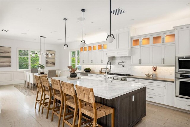 kitchen with an island with sink, stainless steel appliances, decorative backsplash, visible vents, and glass insert cabinets