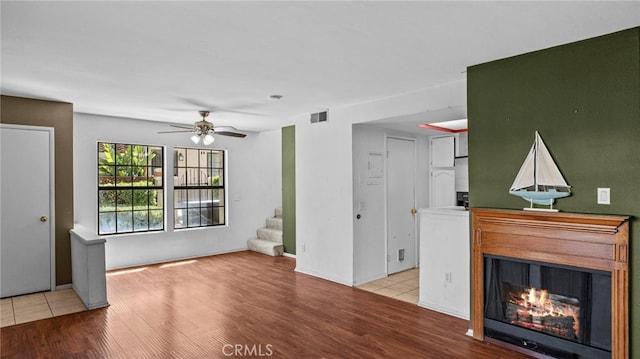 unfurnished living room featuring visible vents, stairs, a glass covered fireplace, light wood-style floors, and a ceiling fan