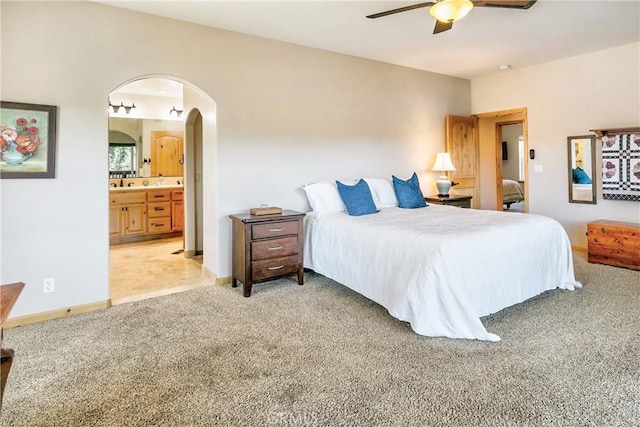 bedroom featuring light colored carpet, ensuite bathroom, baseboards, arched walkways, and a ceiling fan