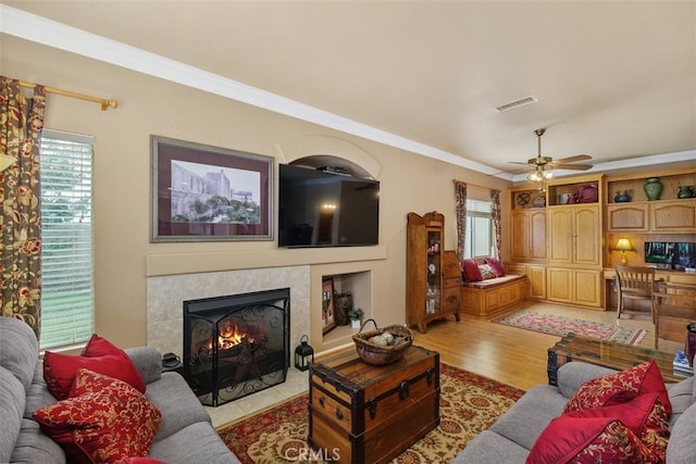 living area featuring light wood finished floors, a tile fireplace, crown molding, a ceiling fan, and visible vents