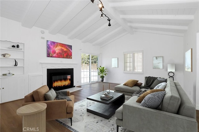living area featuring wood finished floors, vaulted ceiling with beams, rail lighting, a glass covered fireplace, and baseboards