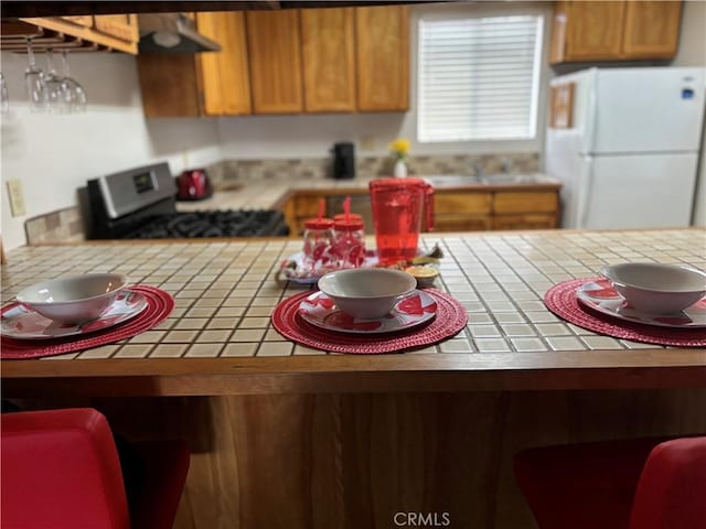 kitchen featuring brown cabinets, freestanding refrigerator, tile countertops, and gas range